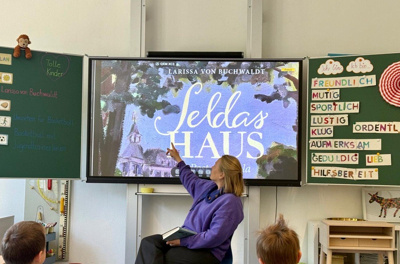Larissa von Buchwaldt liest in einer Grundschulklasse aus Seldas Haus; auf der Tafel ist das Buchcover zu sehen